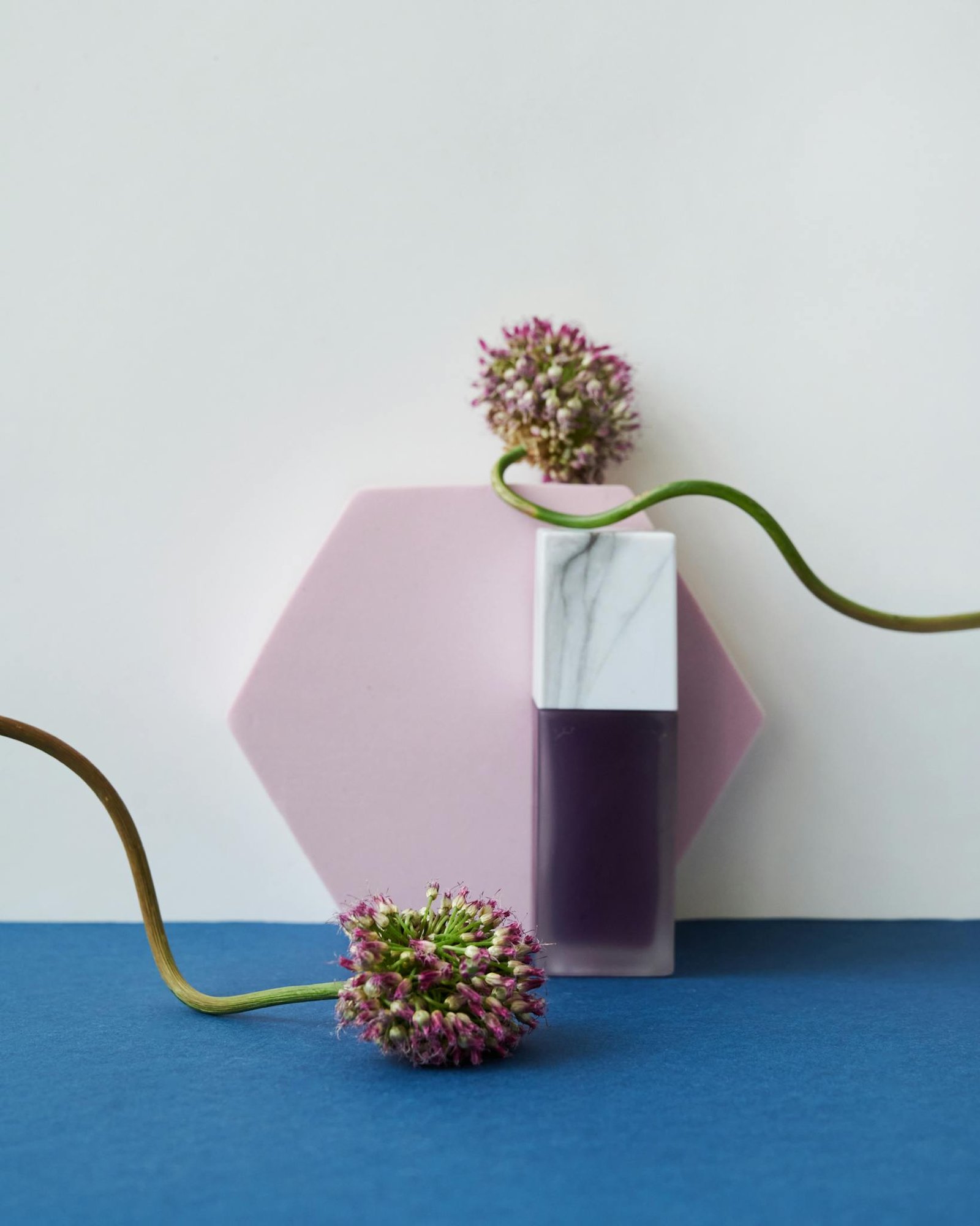 A minimalist still life of a purple perfume bottle and allium flowers on a colorful backdrop.