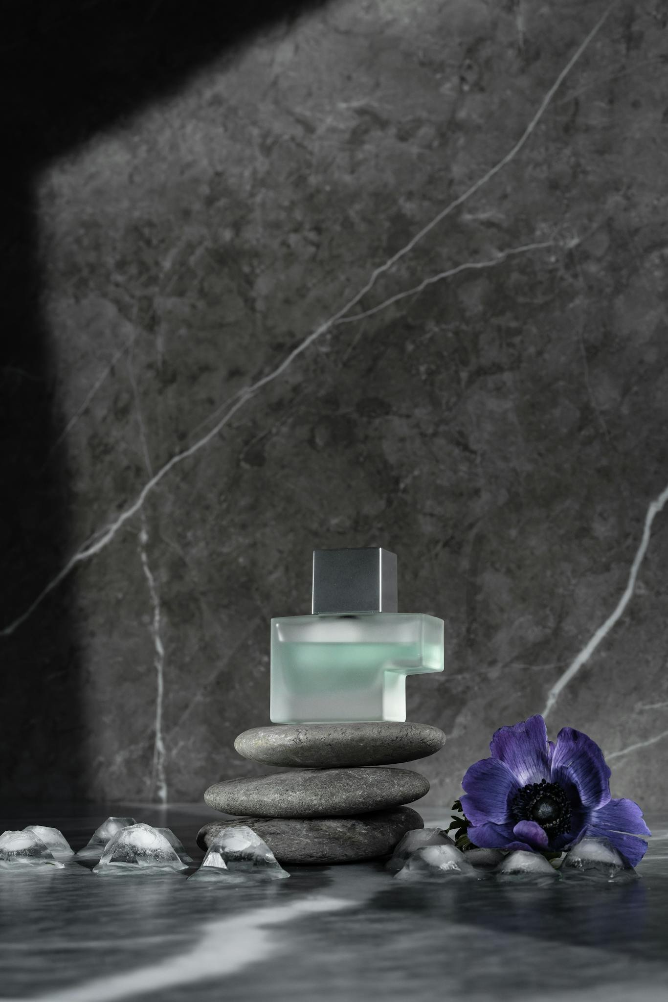 Stylish perfume bottle with purple anemone and balanced stones on gray marble background.